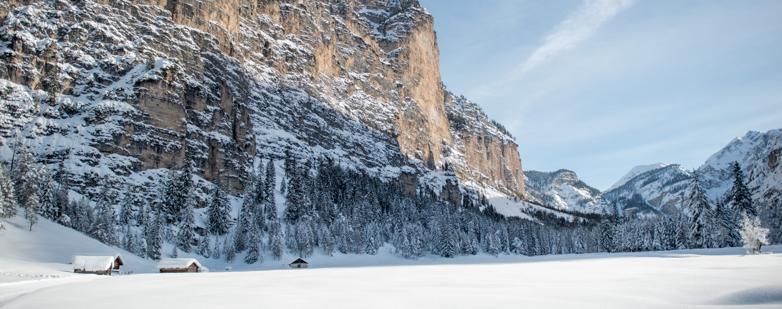 Schneebedeckte Hütten am Fuße eines hohen, felsigen Berges in einer winterlichen Waldlandschaft. - Camping Al Plan