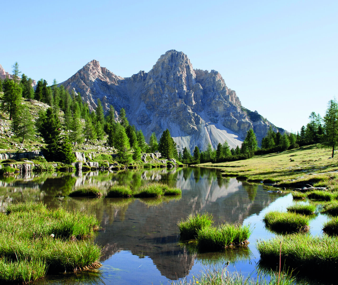 Ein Berg, der sich in einem ruhigen Teich spiegelt, umgeben von grünem Gras und Bäumen unter einem klaren blauen Himmel. - Camping Al Plan