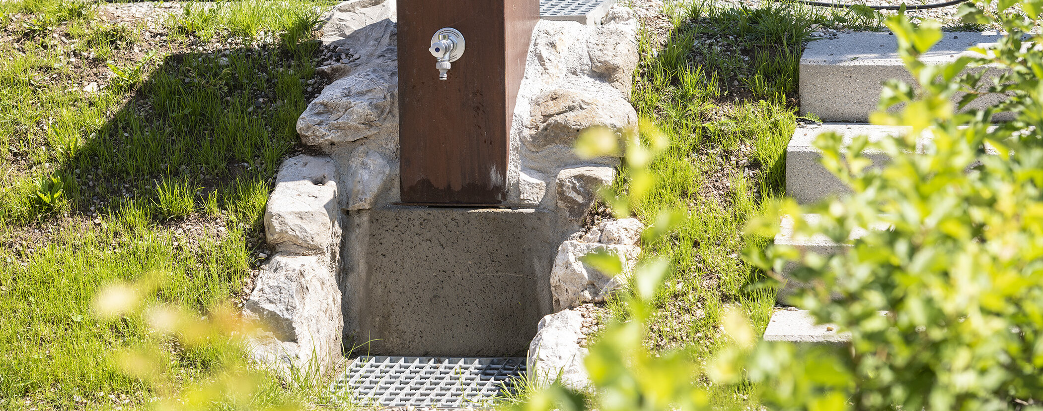 Outdoor water faucet set in rocks, with a drain below and grass and stone steps nearby. - Camping Al Plan