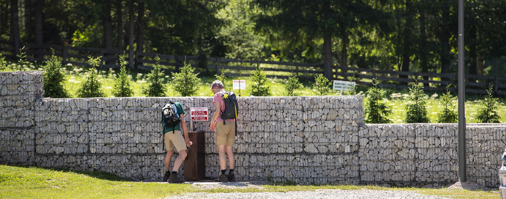 Two hikers with backpacks read signs on a stone wall near trees and a wooden fence on a sunny day. - Camping Al Plan