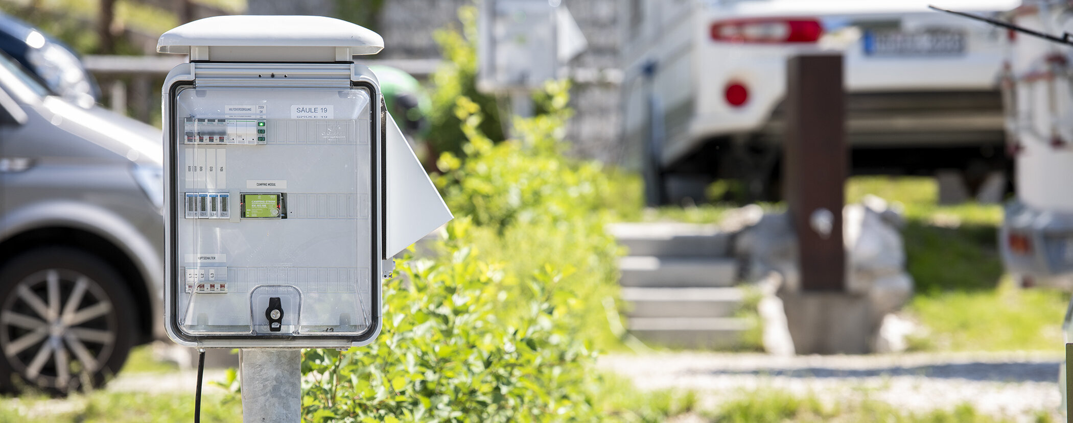 Outdoor electrical control panel near parked cars and greenery, with a white car visible in the background. - Camping Al Plan