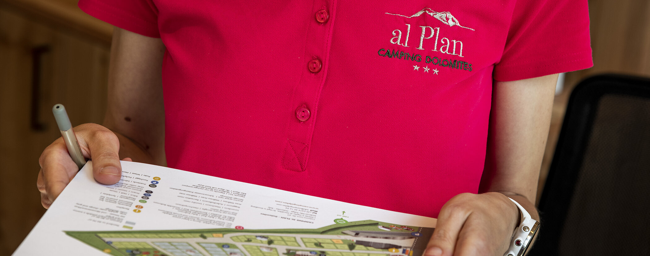 Person in a pink al Plan Camping Dolomiti shirt holding a map and pen. - Camping Al Plan
