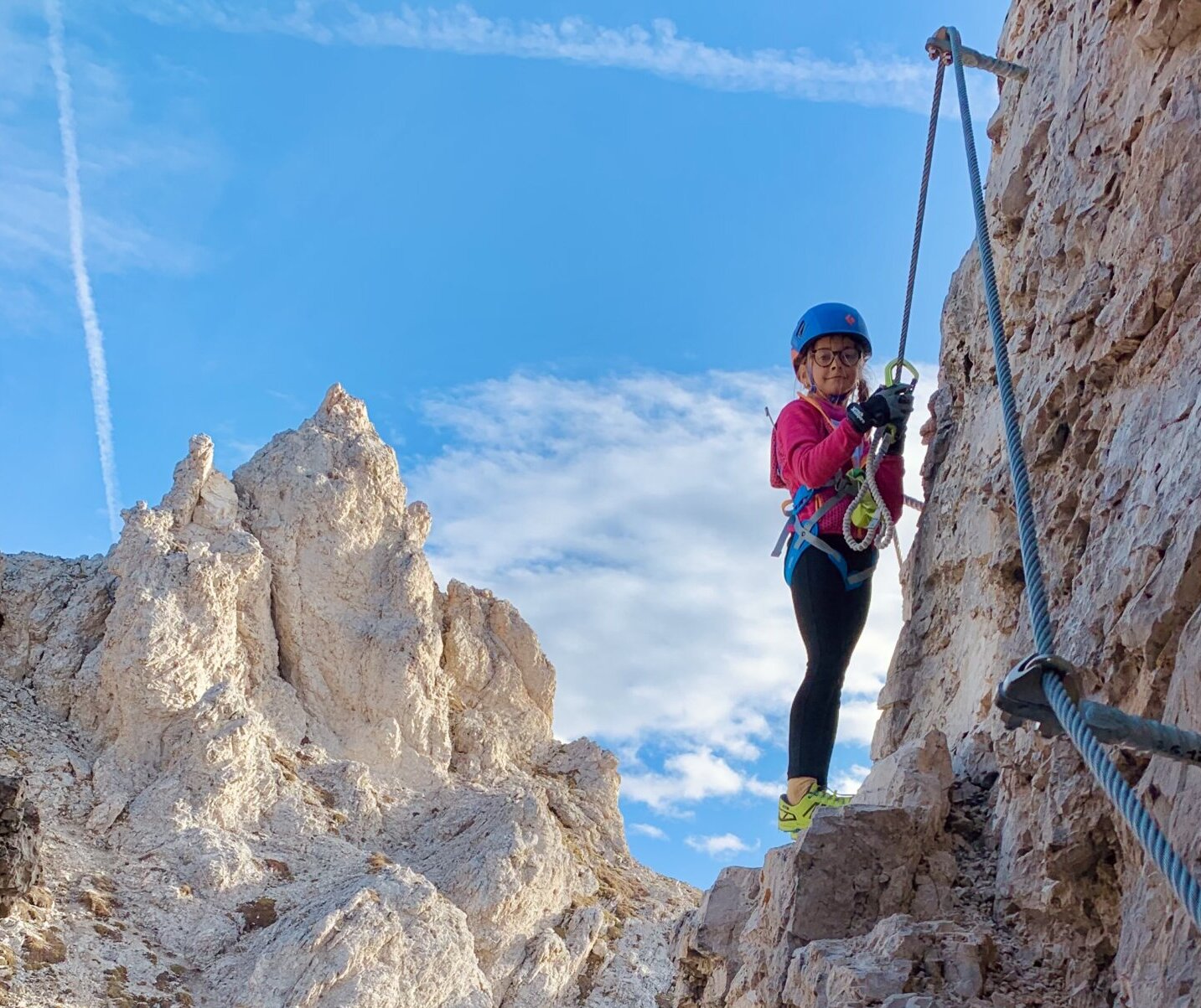Eine Person in Kletterausrüstung erklimmt eine Felswand mit Bergen und blauem Himmel im Hintergrund.