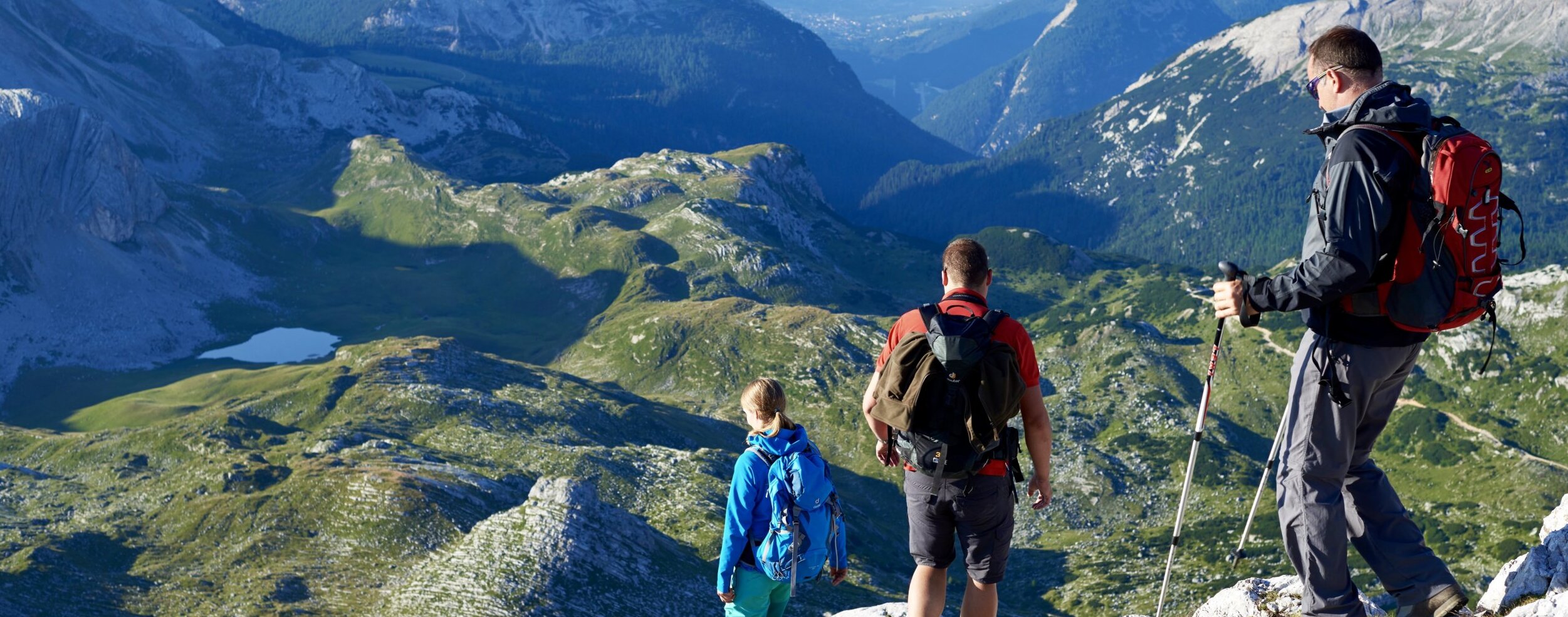 Drei Wanderer mit Rucksäcken stehen auf einem Bergkamm und blicken auf ein grünes Tal und ferne Gipfel. - Camping Al Plan
