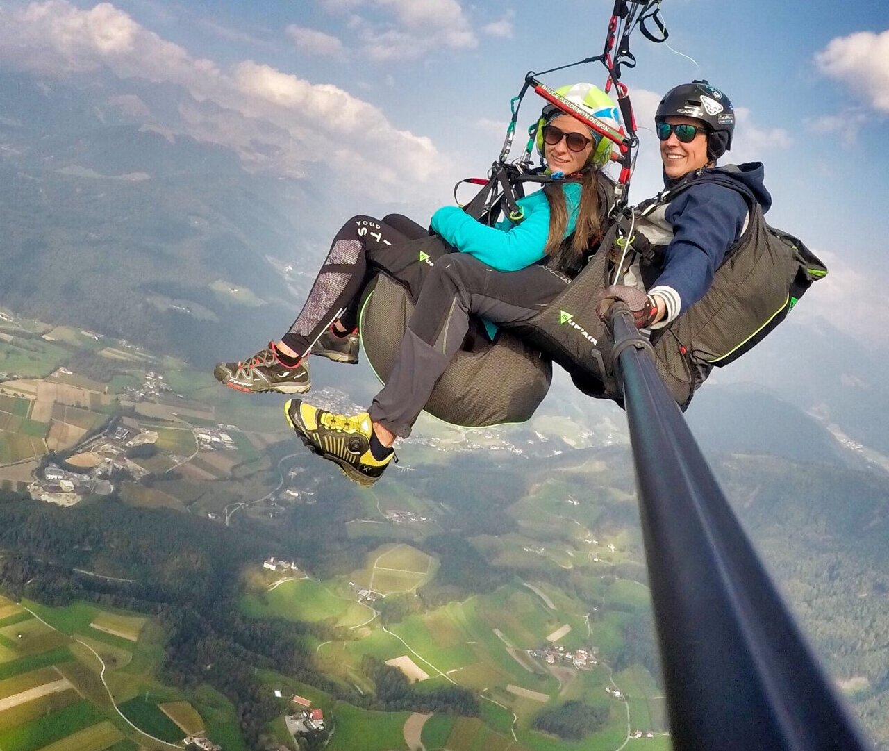 Zwei Personen gleiten mit dem Gleitschirm über eine malerische Landschaft, lächeln und machen ein Selfie mit einem Selfie-Stick. - Camping Al Plan