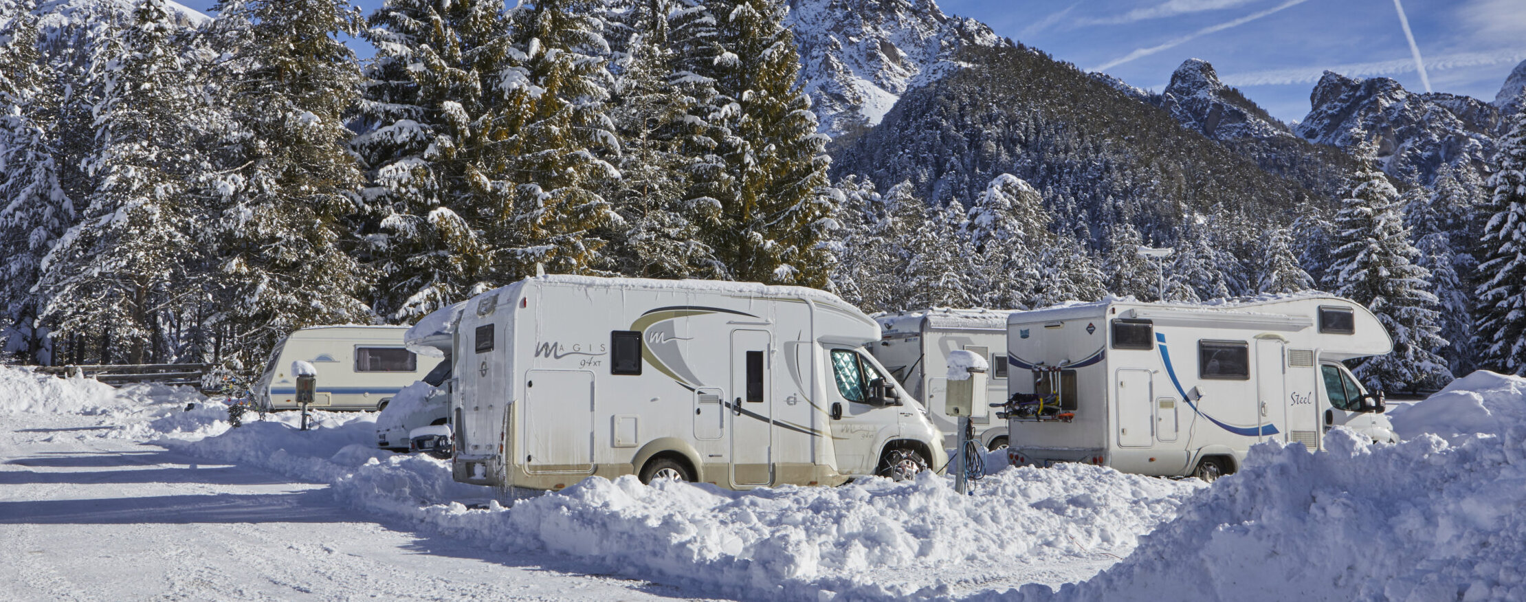 Mehrere Wohnmobile sind in einem verschneiten Gebiet geparkt, das im Winter von Kiefern und Bergen umgeben ist. - Camping Al Plan