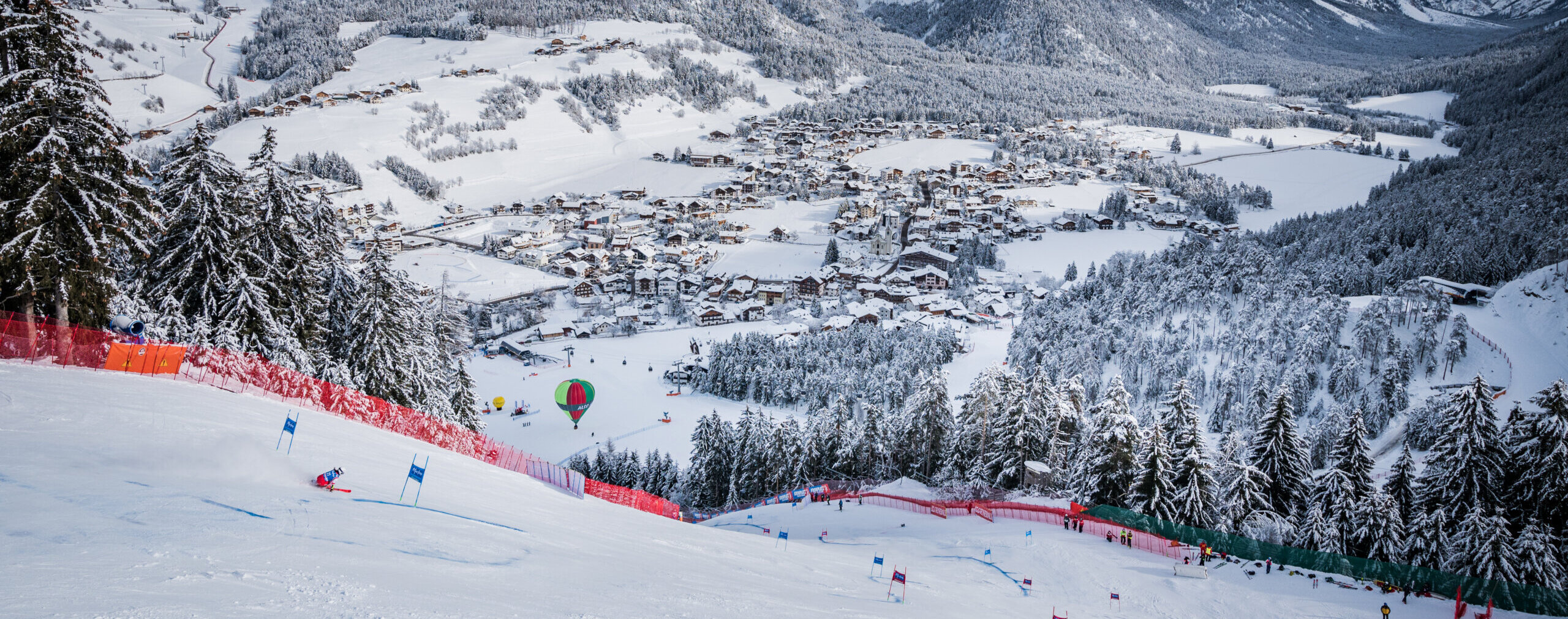 Ein Skifahrer rast auf einem verschneiten Berg in der Nähe eines Dorfes bergab; Heißluftballons schweben über schneebedeckten Bäumen. - Camping Al Plan