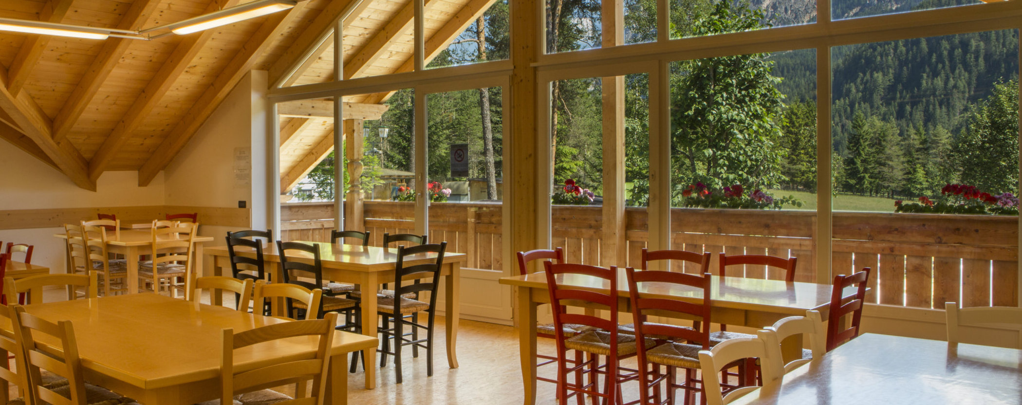 Bright dining room with wooden tables and colorful chairs, large windows overlook a scenic mountain view. - Camping Al Plan