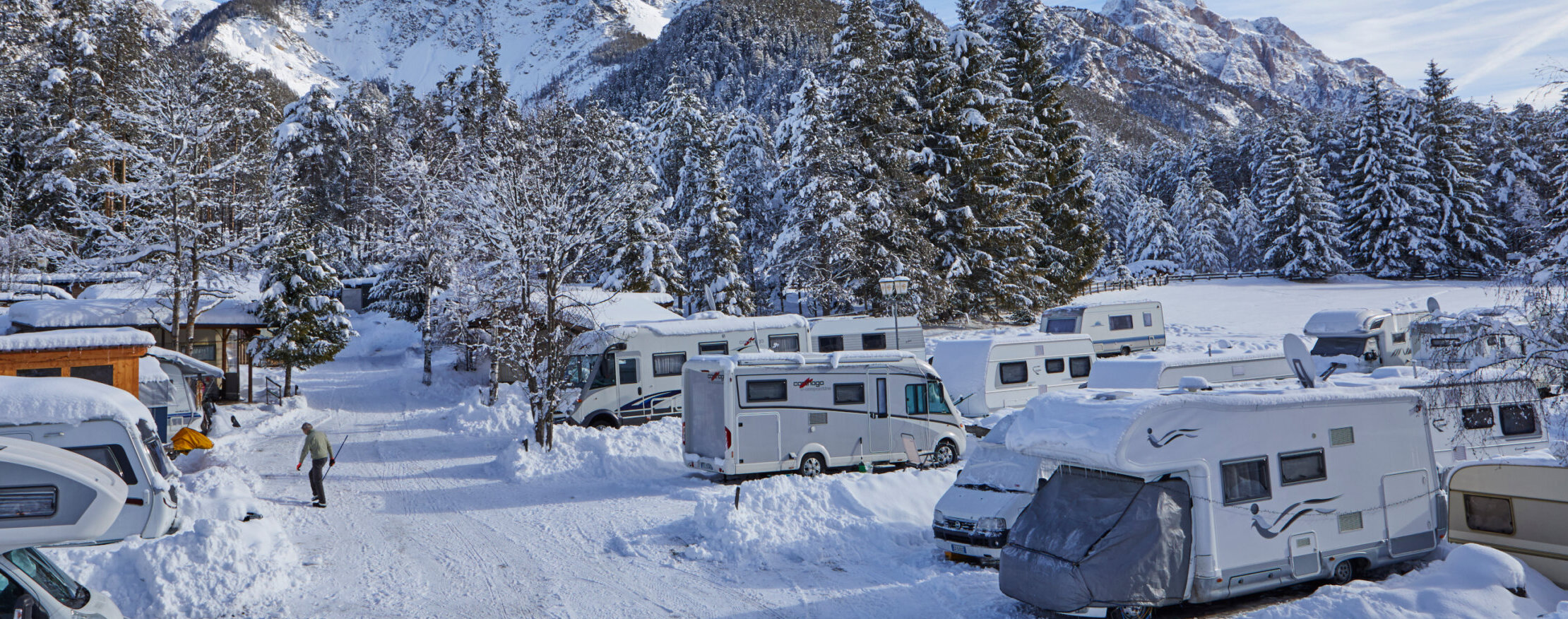 Verschneite Wohnmobile und Tannenbäume auf einem Campingplatz in den Bergen, durch den eine Person an einem Wintertag spaziert. - Camping Al Plan