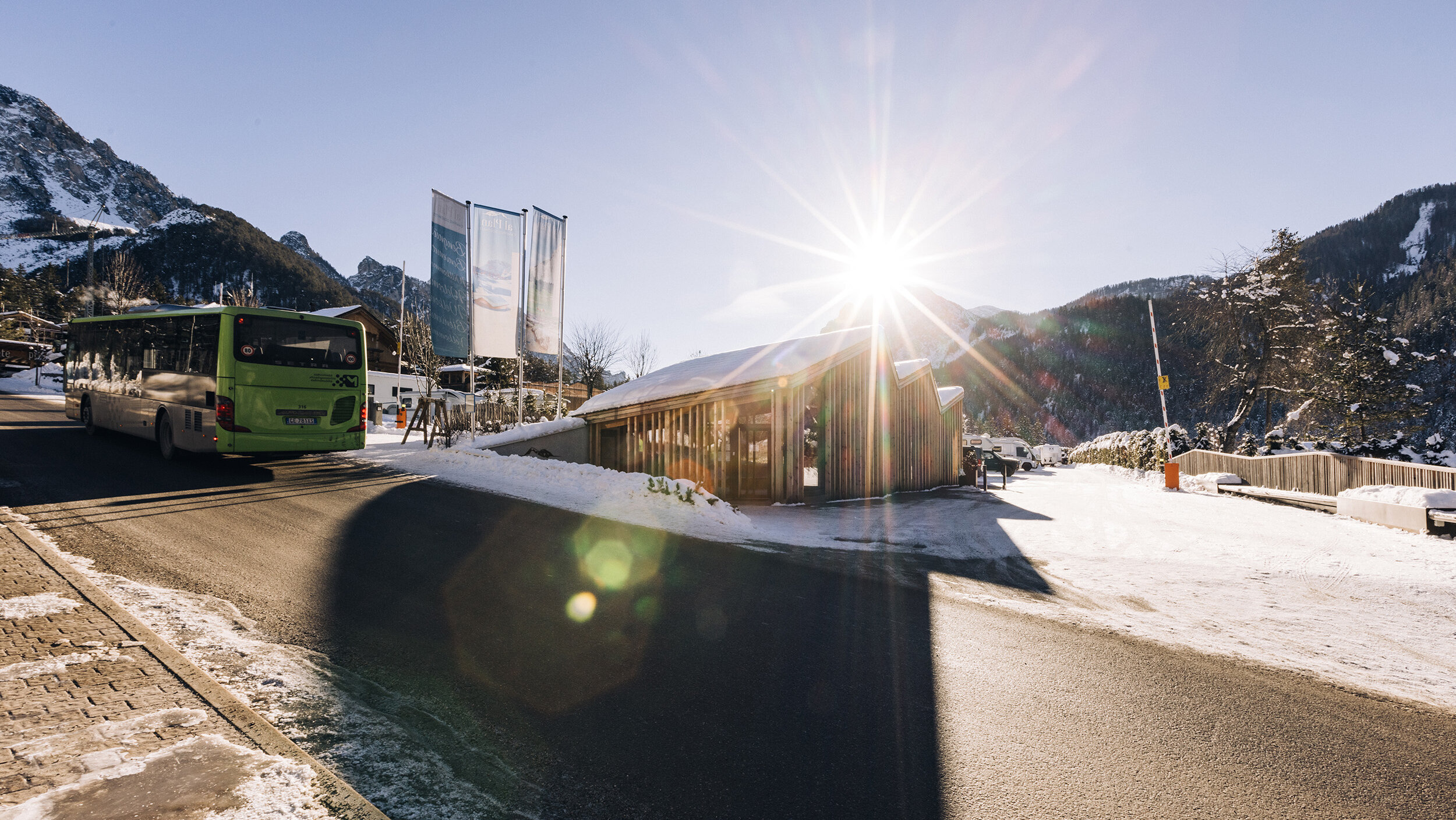 A green bus drives on a sunny, snowy mountain road with a wooden cabin and bright sun in the sky. - Camping Al Plan
