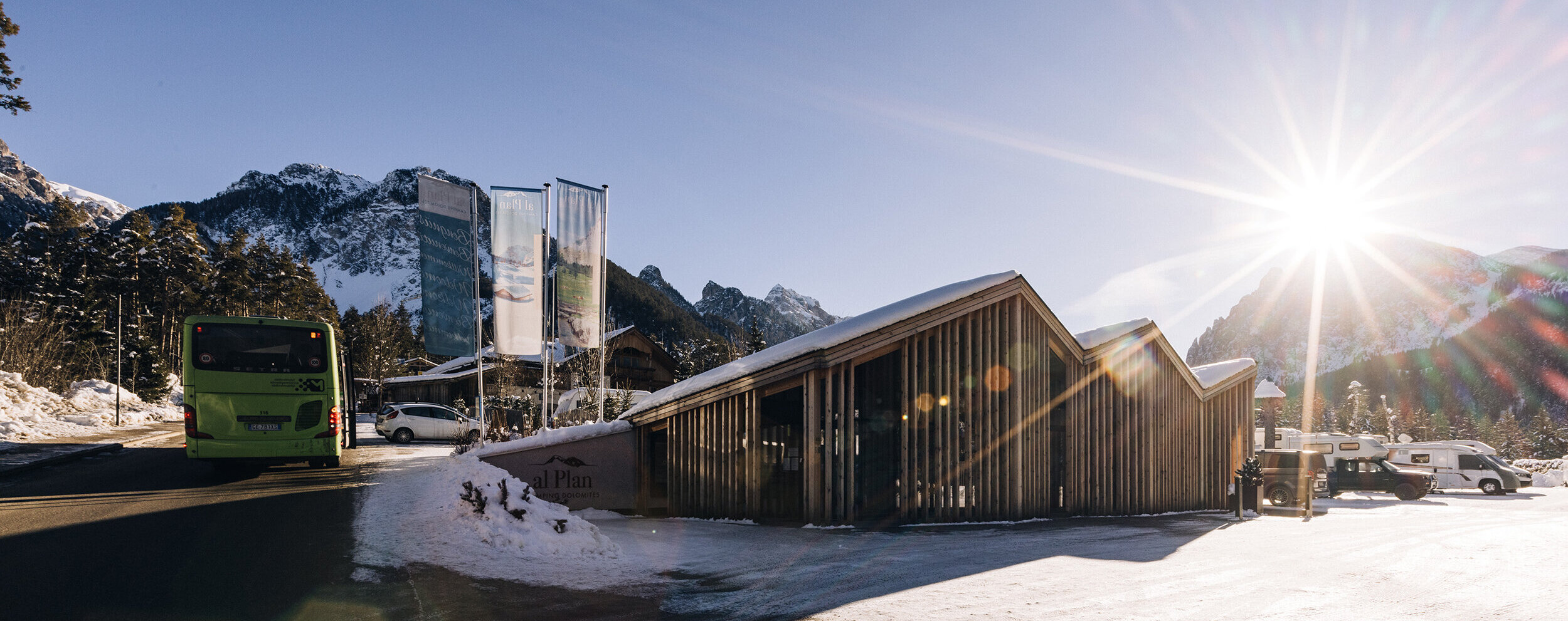 Bei strahlendem Sonnenschein und klarem Himmel fährt ein grüner Bus an verschneiten Hütten und Bergen vorbei. - Camping Al Plan