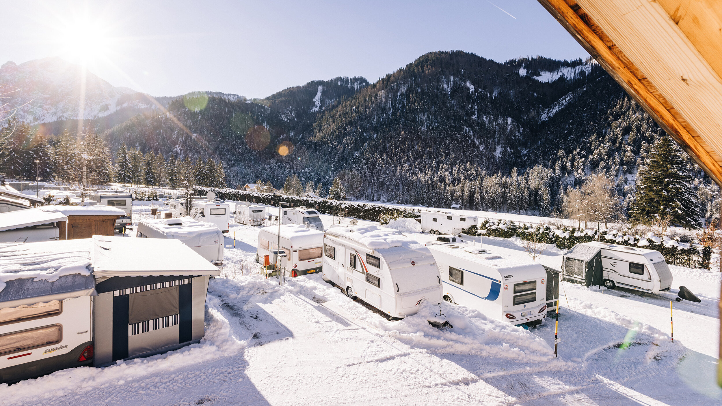 Rows of snow-covered camper vans and cabins in a scenic winter mountain landscape under a bright sun. - Camping Al Plan