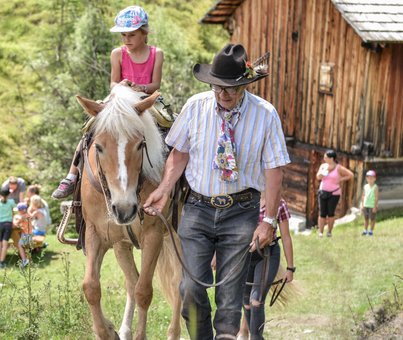 Ein Mann führt ein Pferd, auf dem ein junges Mädchen reitet, in der Nähe eines rustikalen Holzgebäudes im Freien. - Camping Al Plan