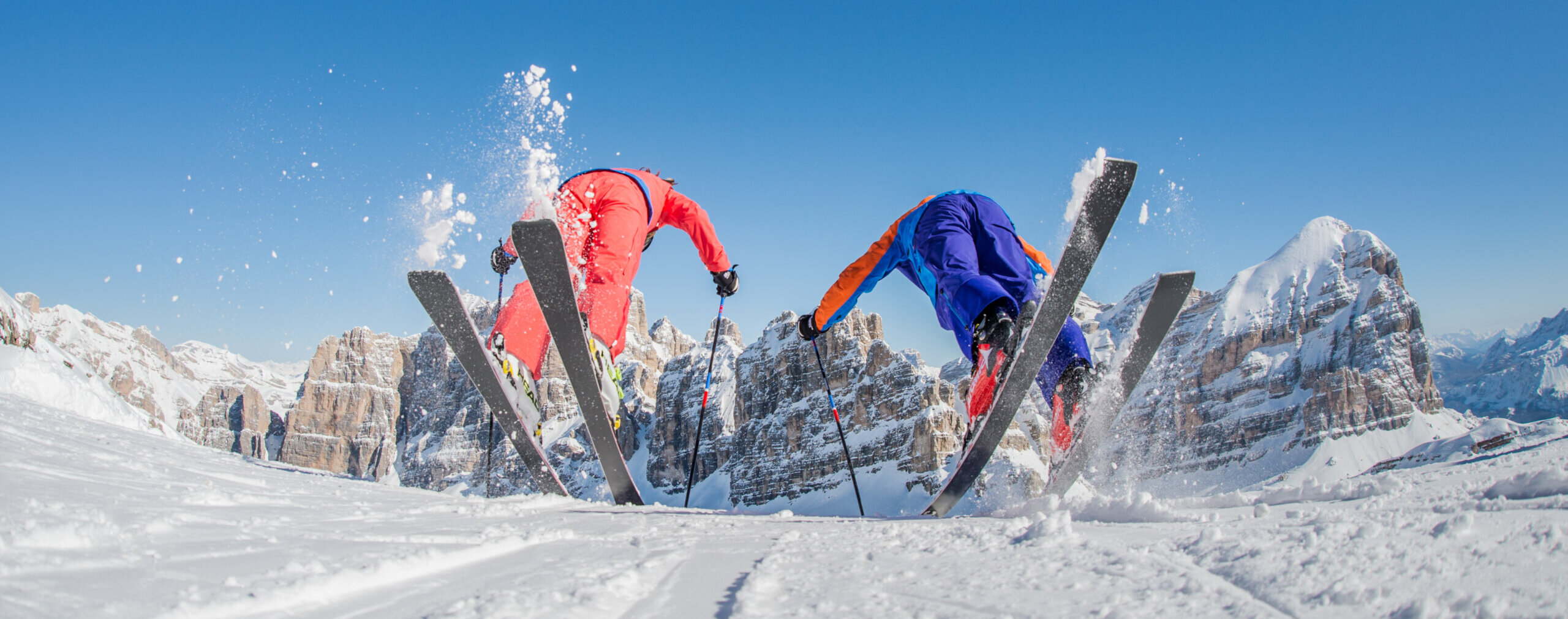 Zwei Skifahrer in bunten Anzügen beim Skifahren auf verschneiten Bergen mit blauem Himmel und schneebedeckten Gipfeln im Hintergrund. - Camping Al Plan